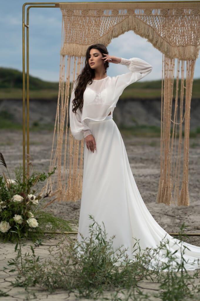 Булчинска рокля с дълъг ръкав "Арабела", елегантна с прозрачни дантелени ръкави и изящен шлейф на фона на природен пейзаж.