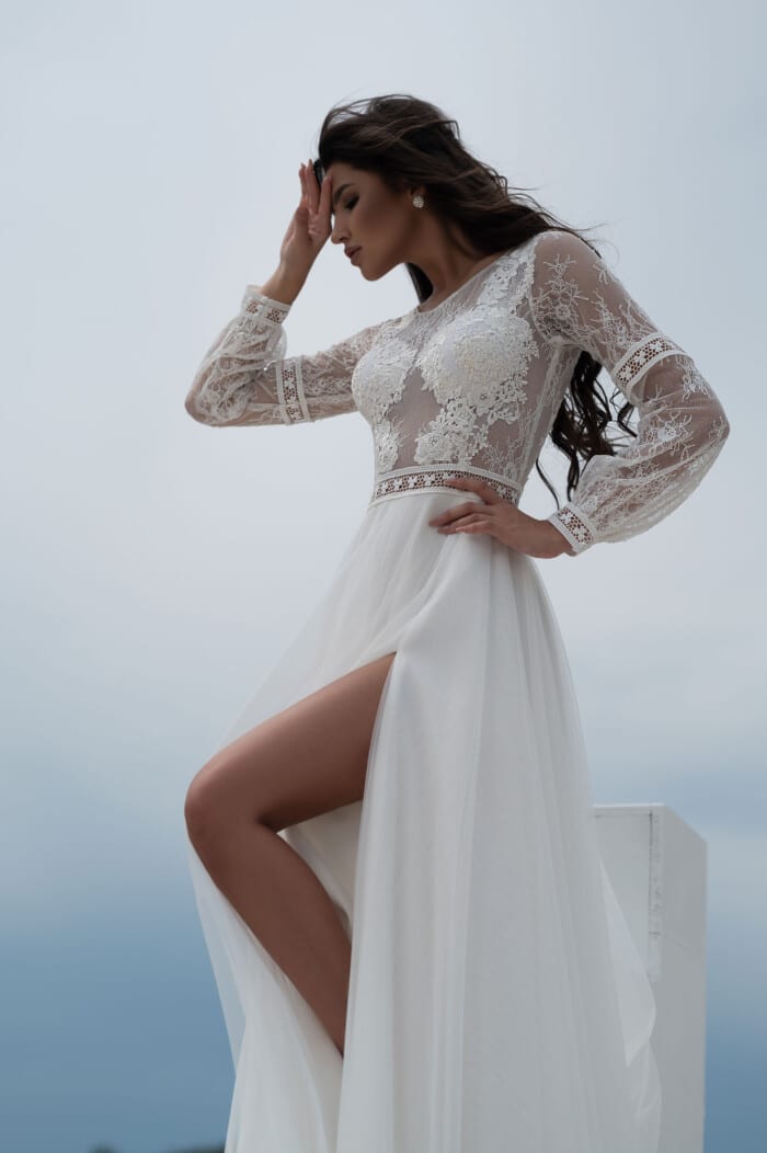 Модел позира със булчинска рокля с дантелени ръкави и елегантна цепка, идеална за съвременната булка