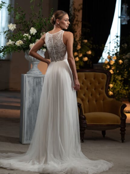 Пемиум булчинска рокля с дълъг шлейф модел Габи, с изящни дантели и апликации, представена в луксозен салон с цветя.