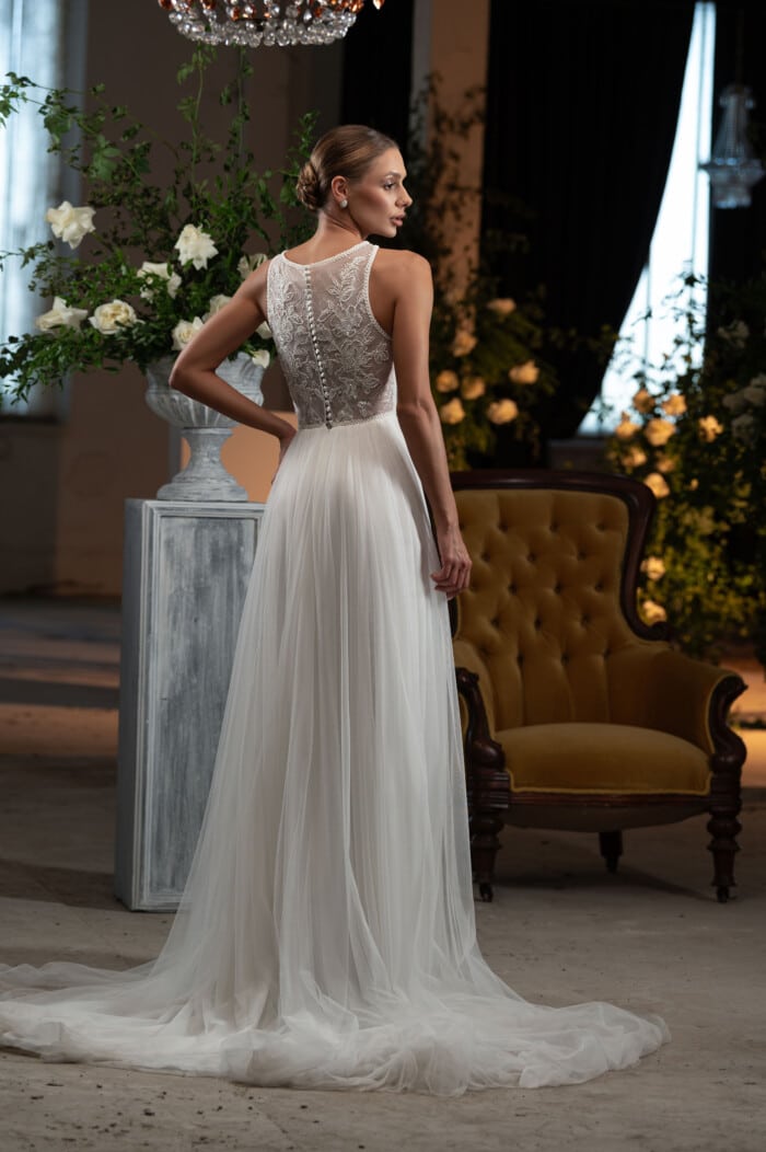 Пемиум булчинска рокля с дълъг шлейф модел Габи, с изящни дантели и апликации, представена в луксозен салон с цветя.