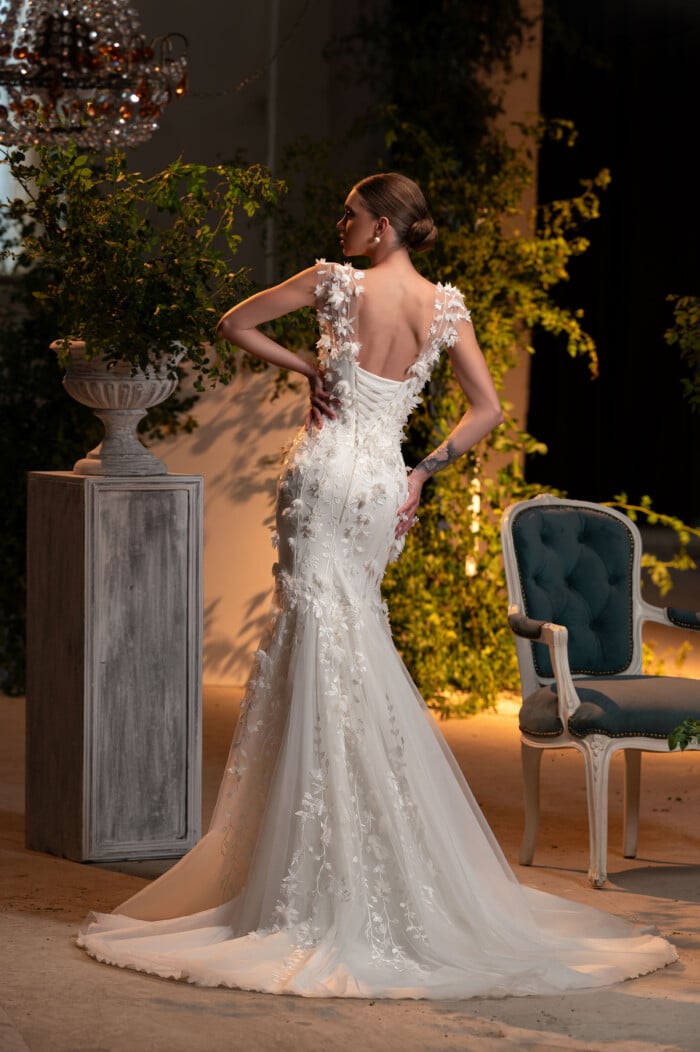 Булчинска рокля модел Мабел, с изящни 3Д апликации, представена в луксозна стая с растения.