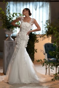 Булчинска рокля с флорални мотиви модел Мабела, с изящни 3Д апликации, представена в луксозна стая с растения.