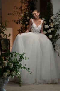 Сватбени тюлени рокли Ашли с деликатни флорални мотиви и пищен шлейф, разположена в елегантен интериор с цветя.