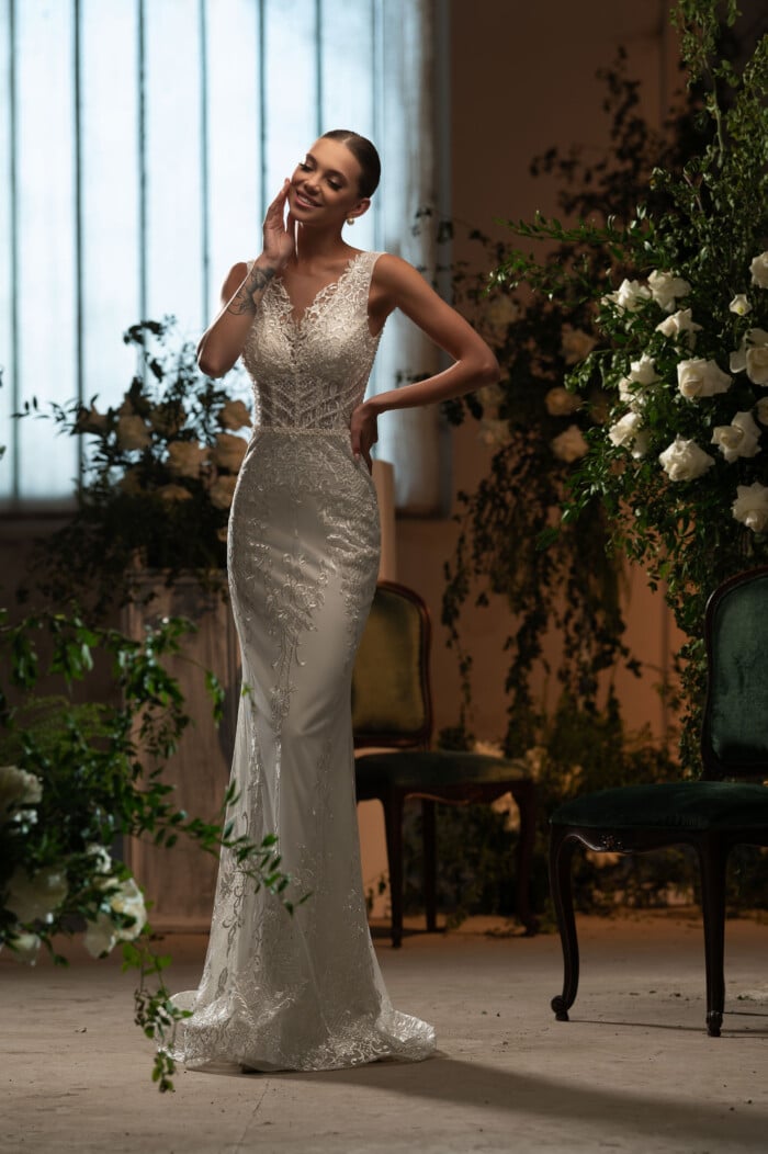 Тясна сватбена рокля по тялото Камерън с елегантни дантелени детайли и стилно V-образно деколте, позирана в романтична обстановка.