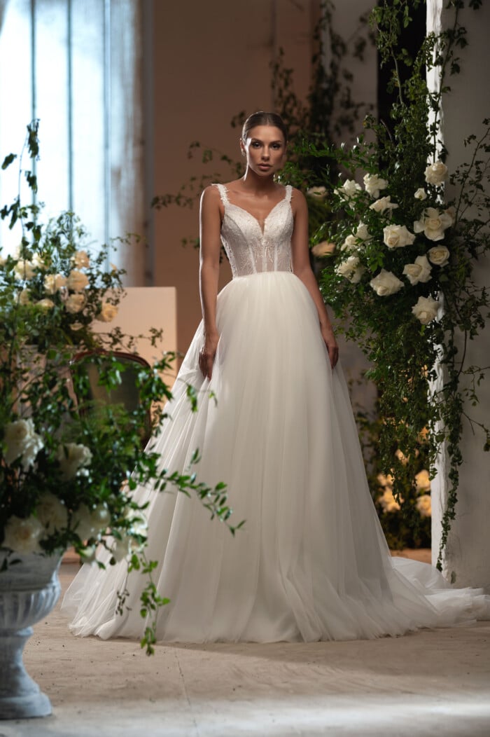 Елегантна булка в дизайнерска сватбена рокля Кармелина с деликатна дантела и V-образно деколте, застанала в романтична сватбена обстановка.