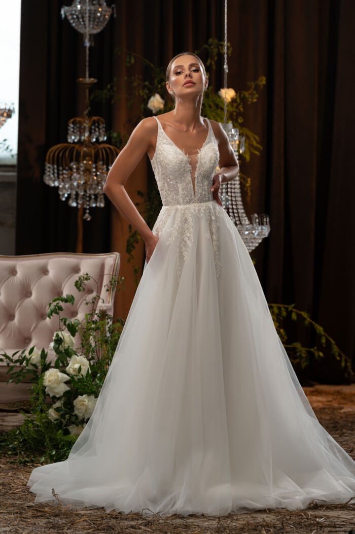 Булка в сватбена рокля с дантела - Милен с детайли и V-образно деколте, стои пред луксозен интериор с полилеи с кристали.