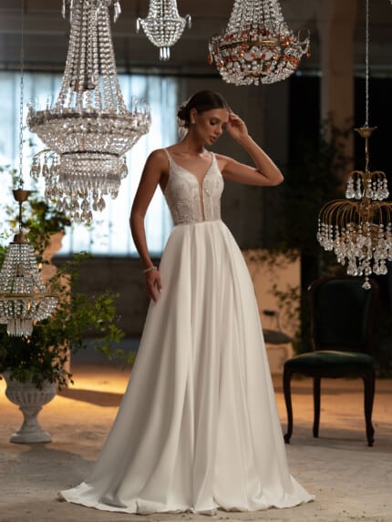 Елегантни булчински рокли Оливия - булка облечена в елегантна сватбена рокля с изискани бродерии, позираща под светлината на кристални полилеи.