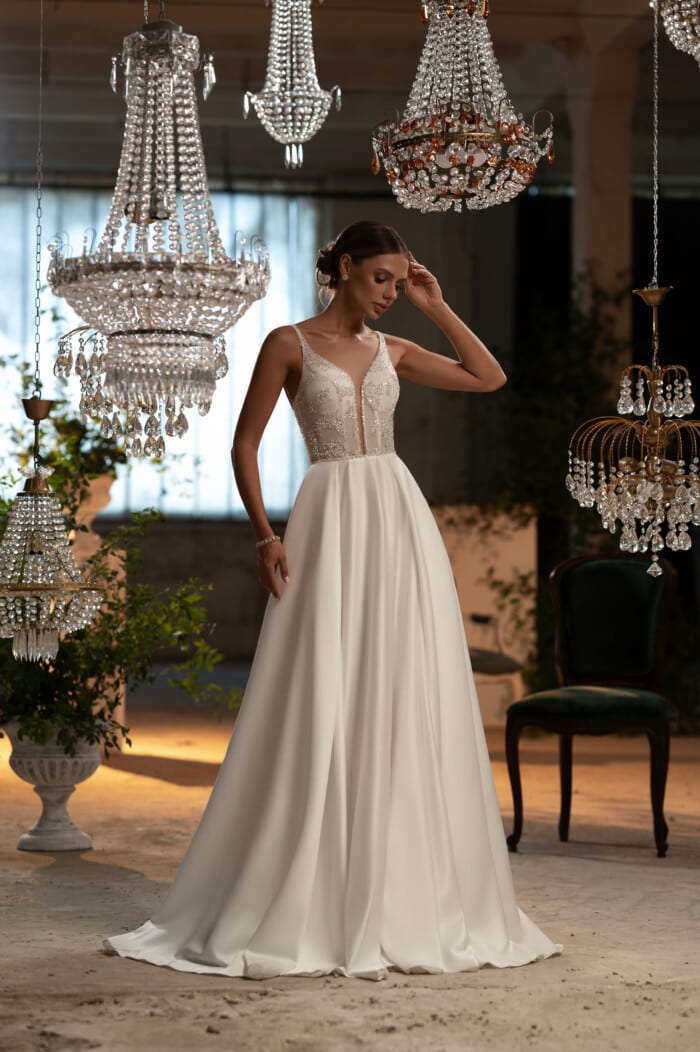 Елегантни булчински рокли Оливия - булка облечена в елегантна сватбена рокля с изискани бродерии, позираща под светлината на кристални полилеи.