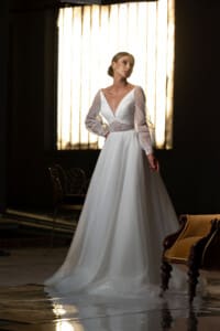 Изящна сватбена рокля "Силвия" с дантелени ръкави и елегантно деколте, позирана пред светлинни ленти.
