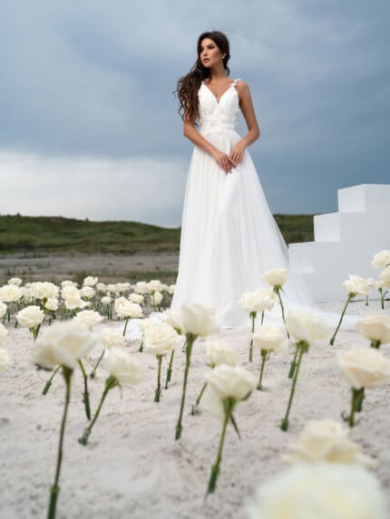 Модел позира със сватбена рокля с гол гръб Сю с флорални акценти, разкриваща елегантна А-линия и дълъг шлейф