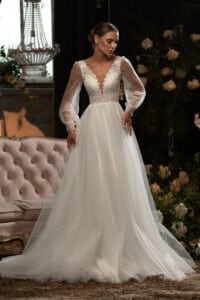Сватбена рокля с дълъг прозрачен ръкав и елегантна бродерия, позира в луксозен салон.