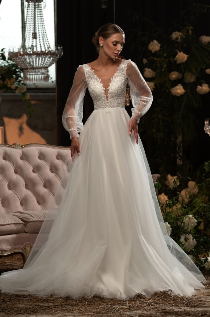 Сватбена рокля с дълъг прозрачен ръкав и елегантна бродерия, позира в луксозен салон.