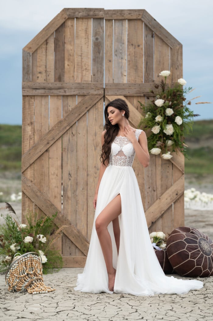 Ръчно изработена сватбена рокля с дълбока цепка Тори с романтично излъчване, позиционирана пред дървена порта